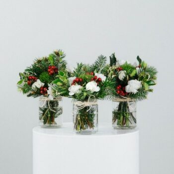three small glass jars decorated with natural twine filled with fresh green spruce red berries and white flowers